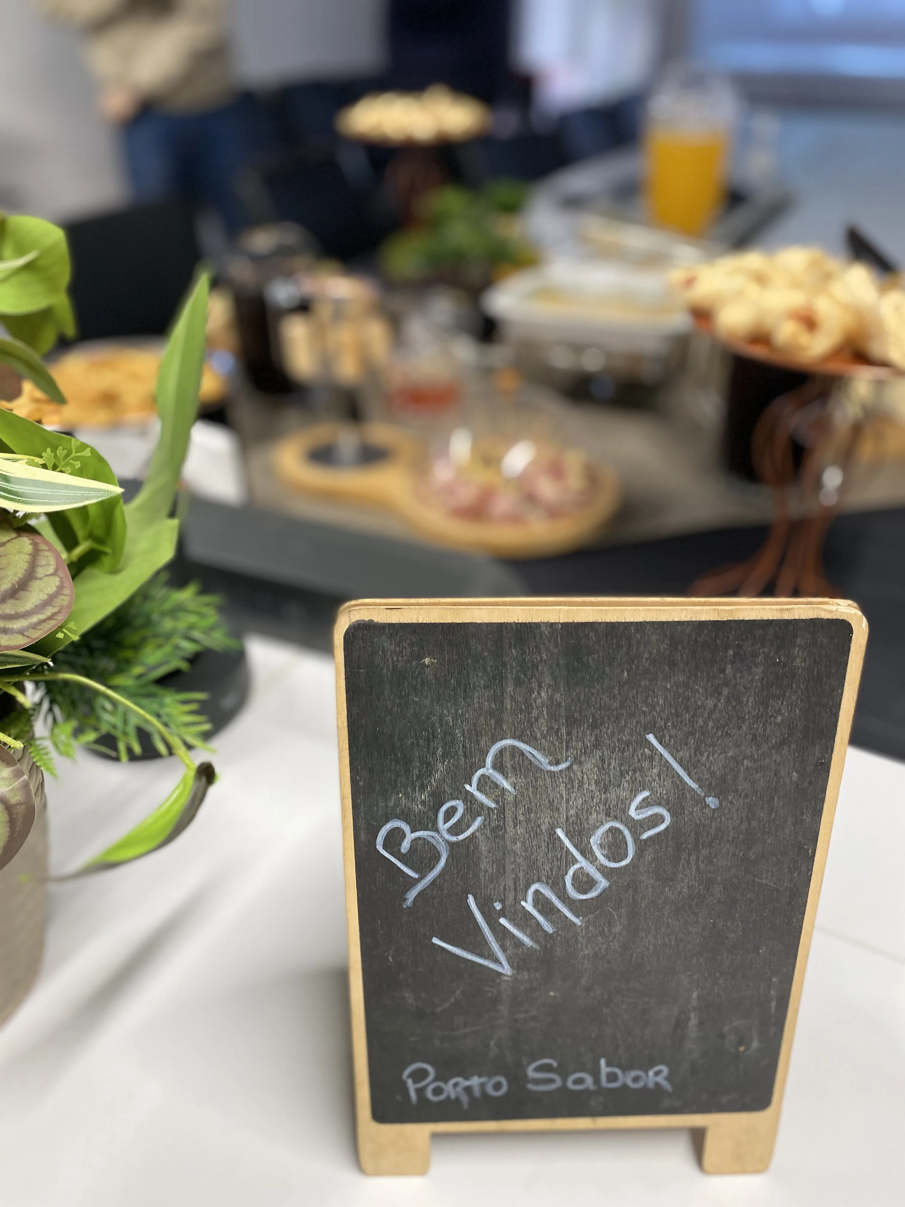 Placa de bem-vindos em mesa de evento Porto Sabor com doces e bebidas ao fundo.